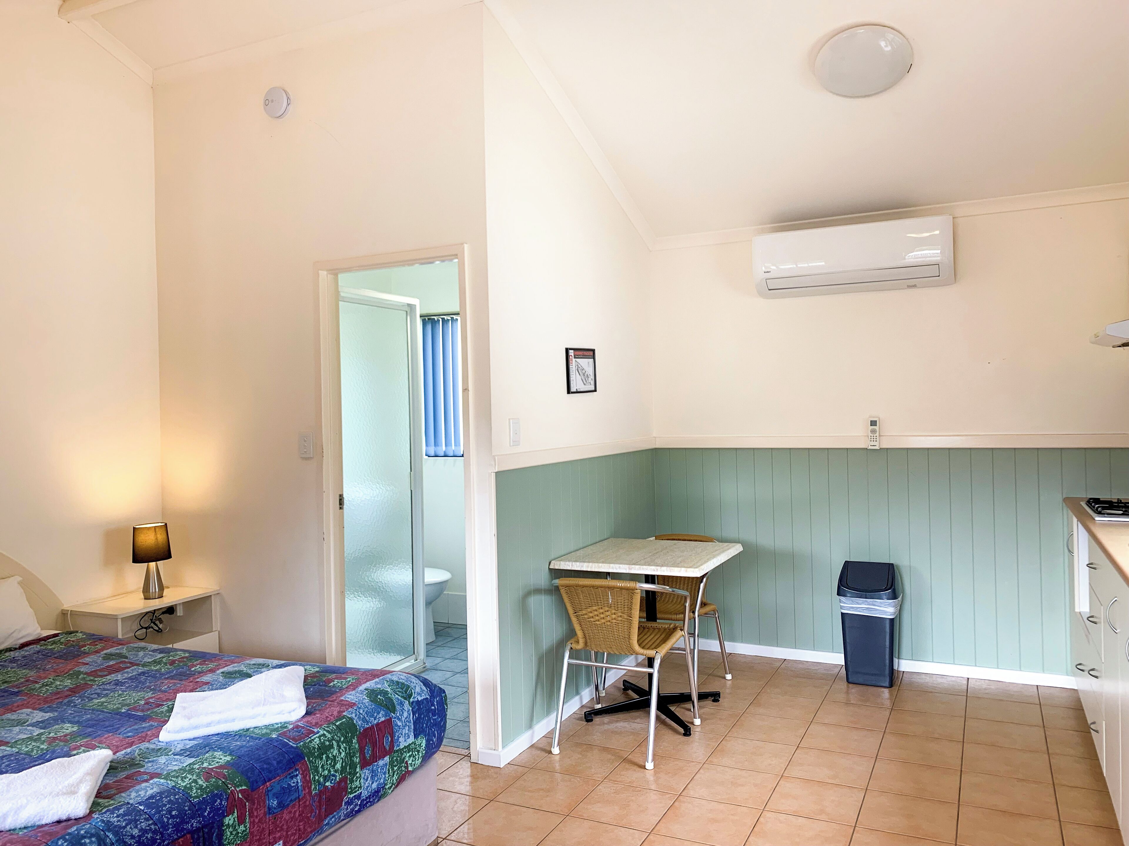 A compact hotel room with a bed, a small desk, a chair, a trash bin, and an air conditioning unit.
