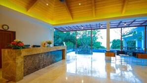 A reception area with a marble counter, wooden ceiling, and large windows offering a view of greenery outside. - Crystal Wild Resort Panwa Phuket (Wichit)