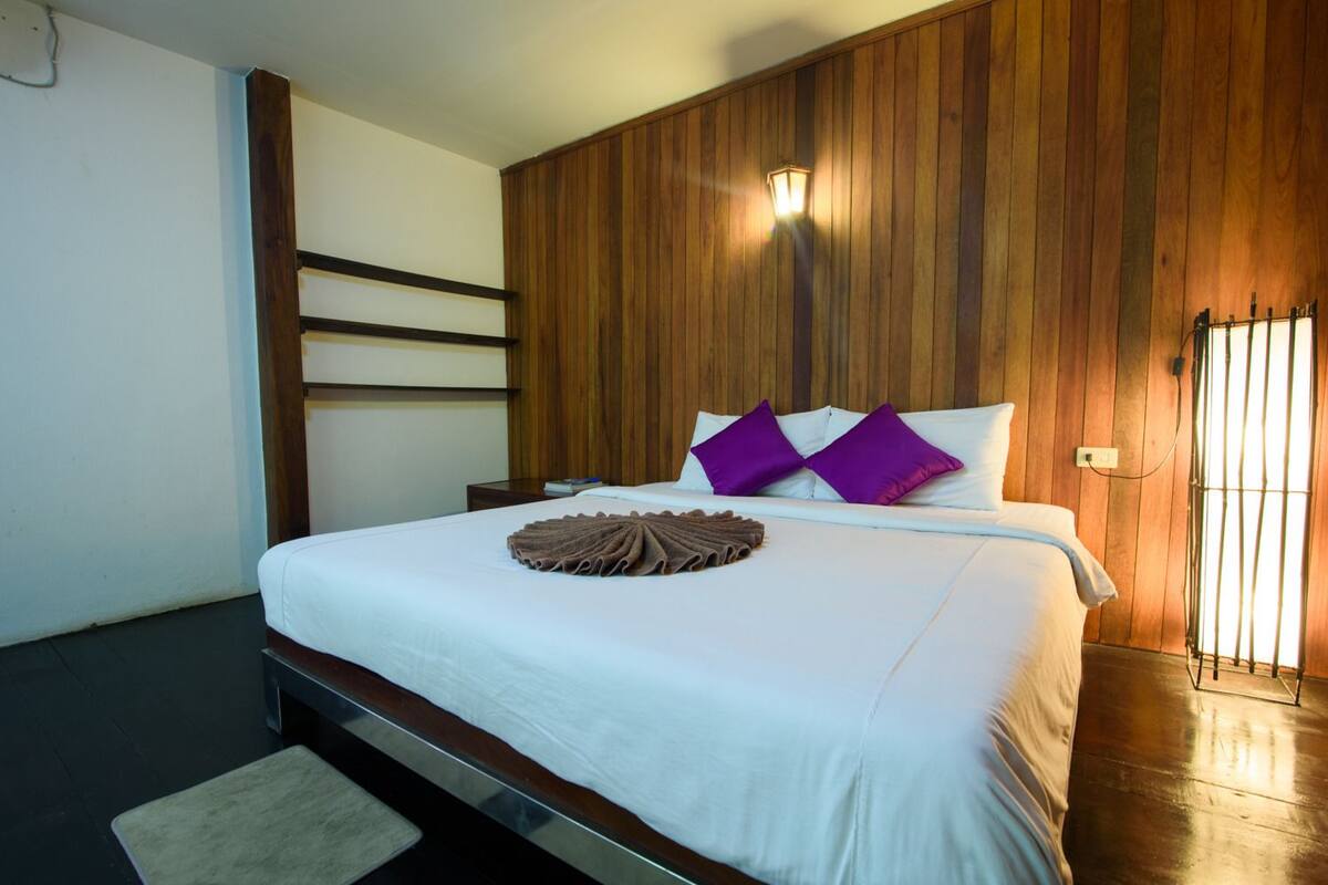 A hotel room with a wooden paneled headboard, a bed with white bedding and purple pillows, a bedside table with a book, and a nightstand with a lamp.