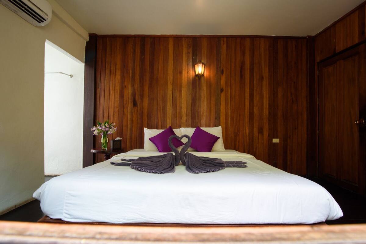 A hotel room with a large bed, wooden panel walls, a nightstand with a vase of flowers, and a wall-mounted lamp.