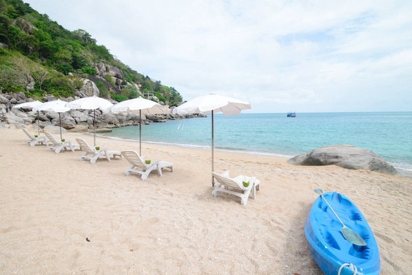 A beach with white lounge chairs and umbrellas, a blue kayak, and a rocky hillside in the background.