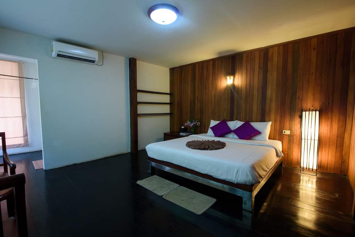 A bedroom with a wooden panel wall, a bed with white bedding and purple pillows, a bedside table with a lamp, and a window with curtains.