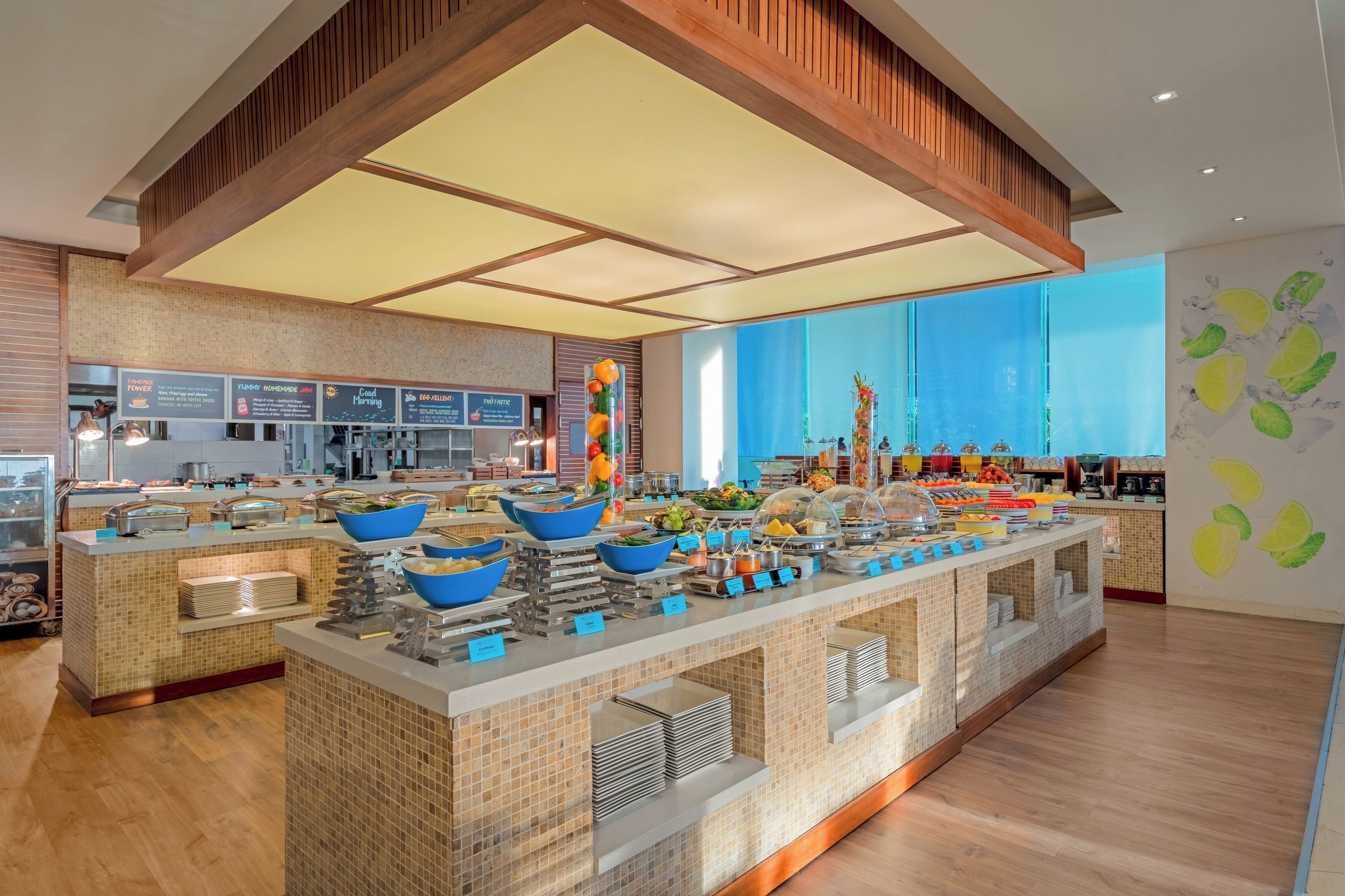 A modern hotel buffet with multiple food stations, a large blue wall mural, and a wooden ceiling.