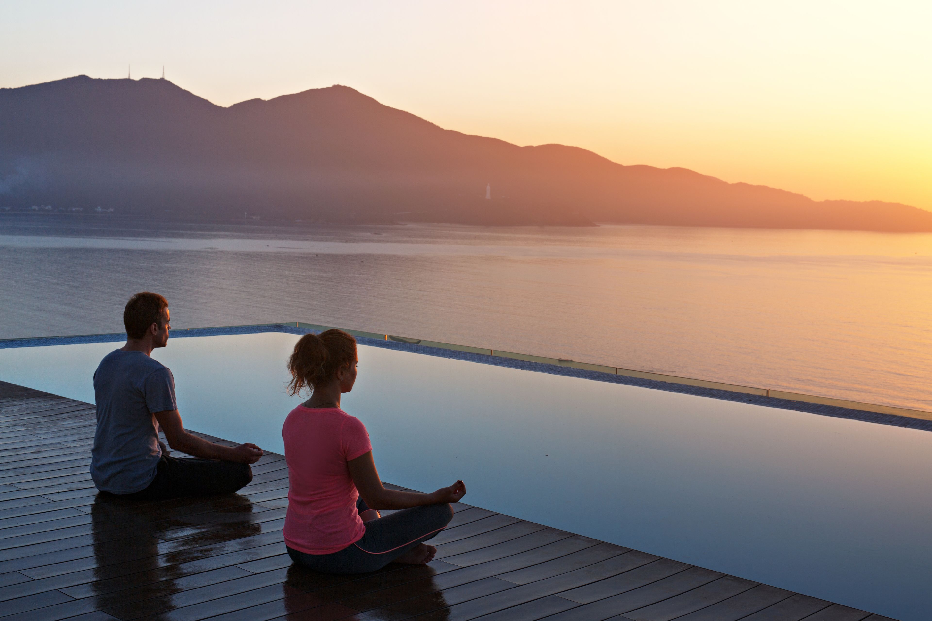 Two people meditating on a wooden deck overlooking a calm body of water with mountains in the background.