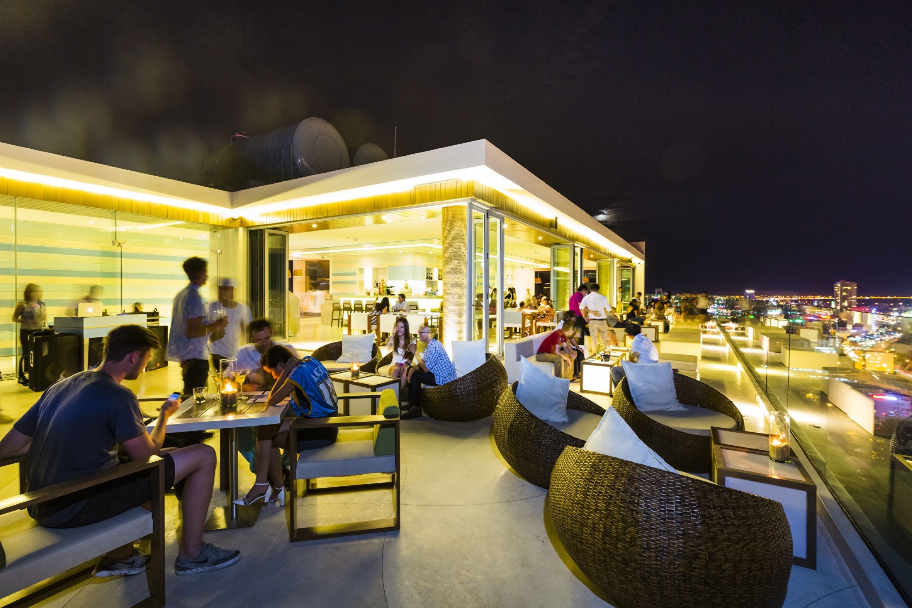An outdoor seating area with wicker furniture and tables, a bar area with people, and a view of the cityscape at night.