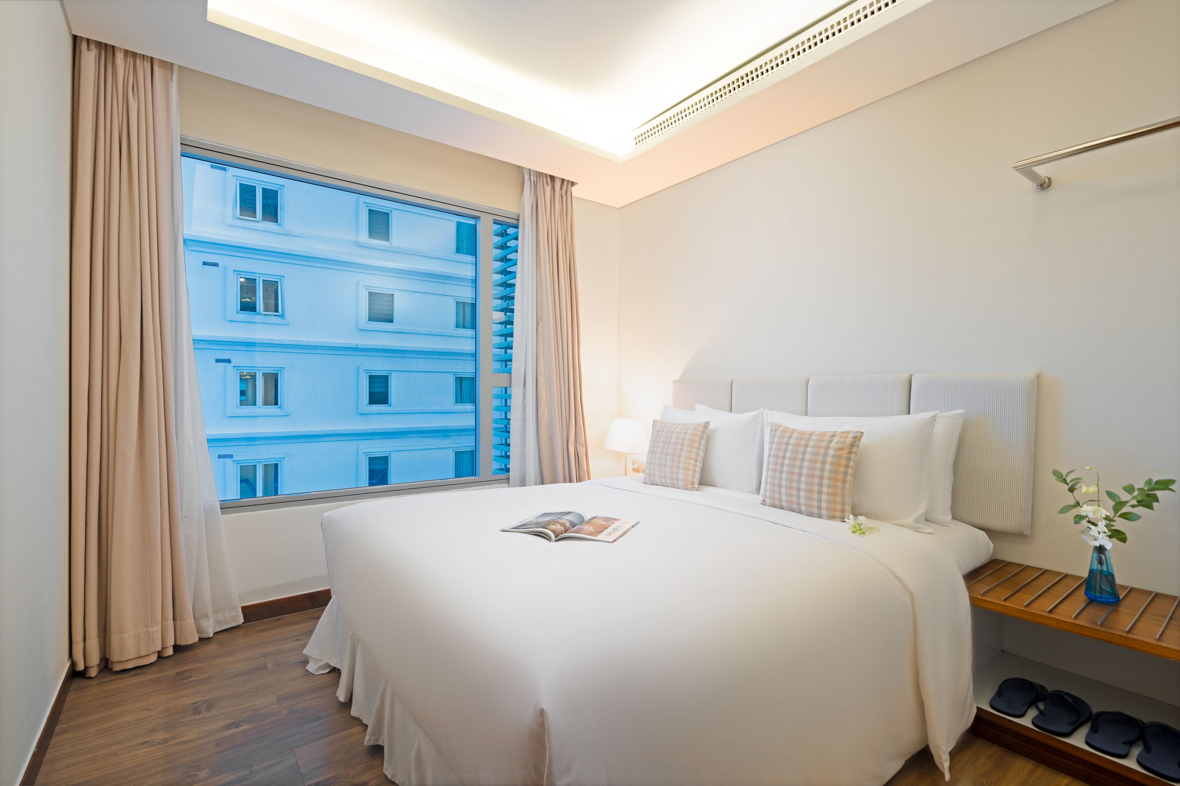 A hotel room with a large bed, a window with curtains, a bedside table with a lamp, and a view of a building outside.