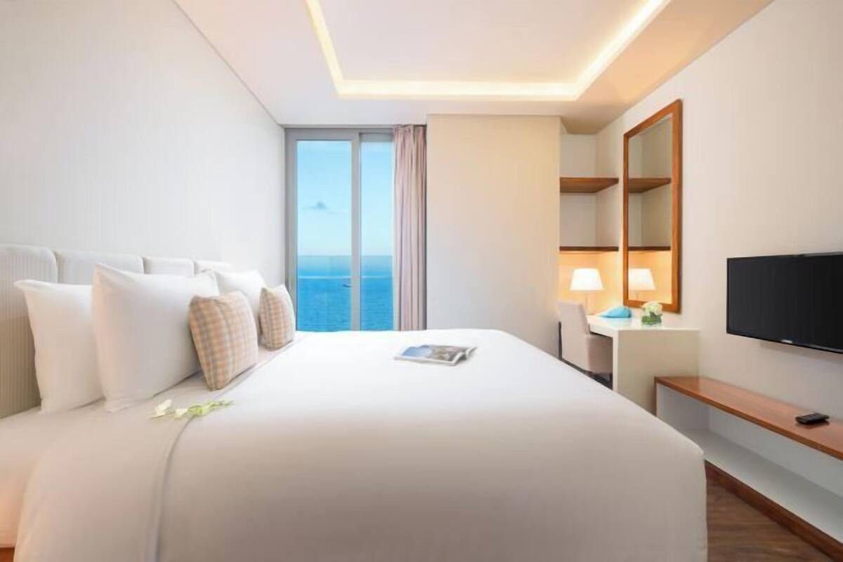 Une chambre d’hôtel avec un grand lit, une télévision, une table de chevet avec une lampe et une vue sur l’océan par la fenêtre.