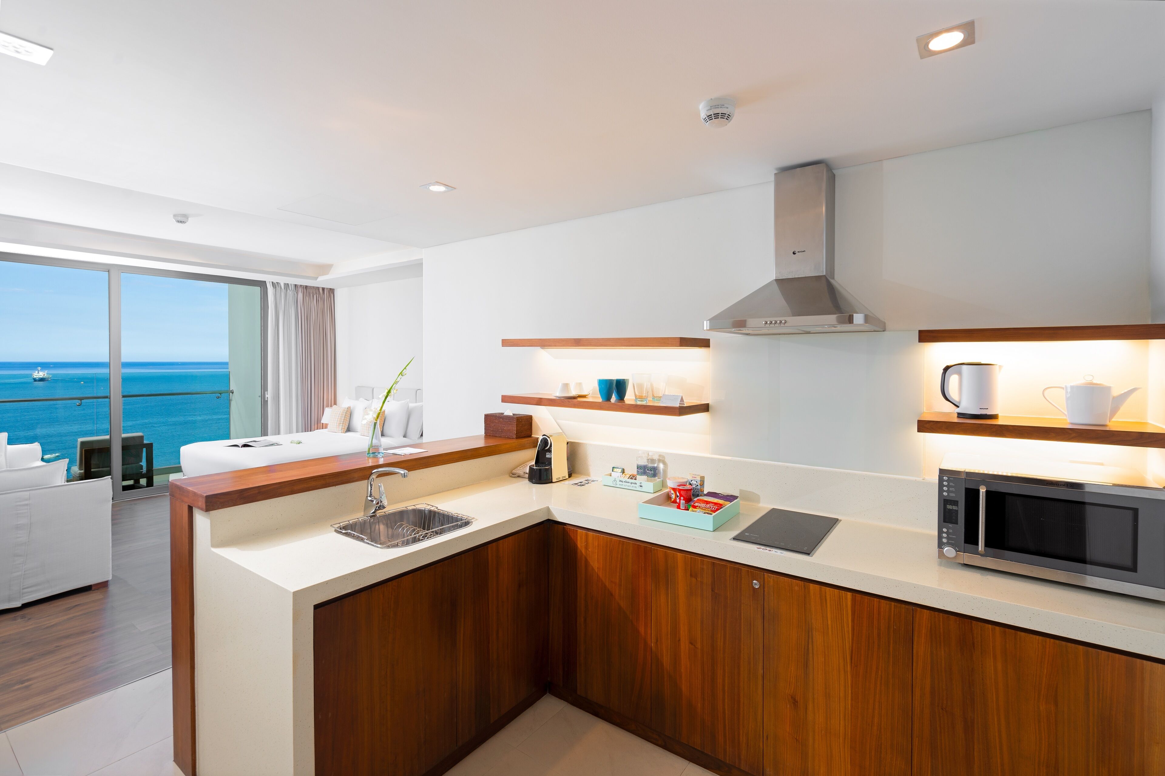 A modern kitchen with a built-in microwave, a sink, and wooden cabinets. There is a breakfast bar with a coffee maker and a view of the ocean through large windows.