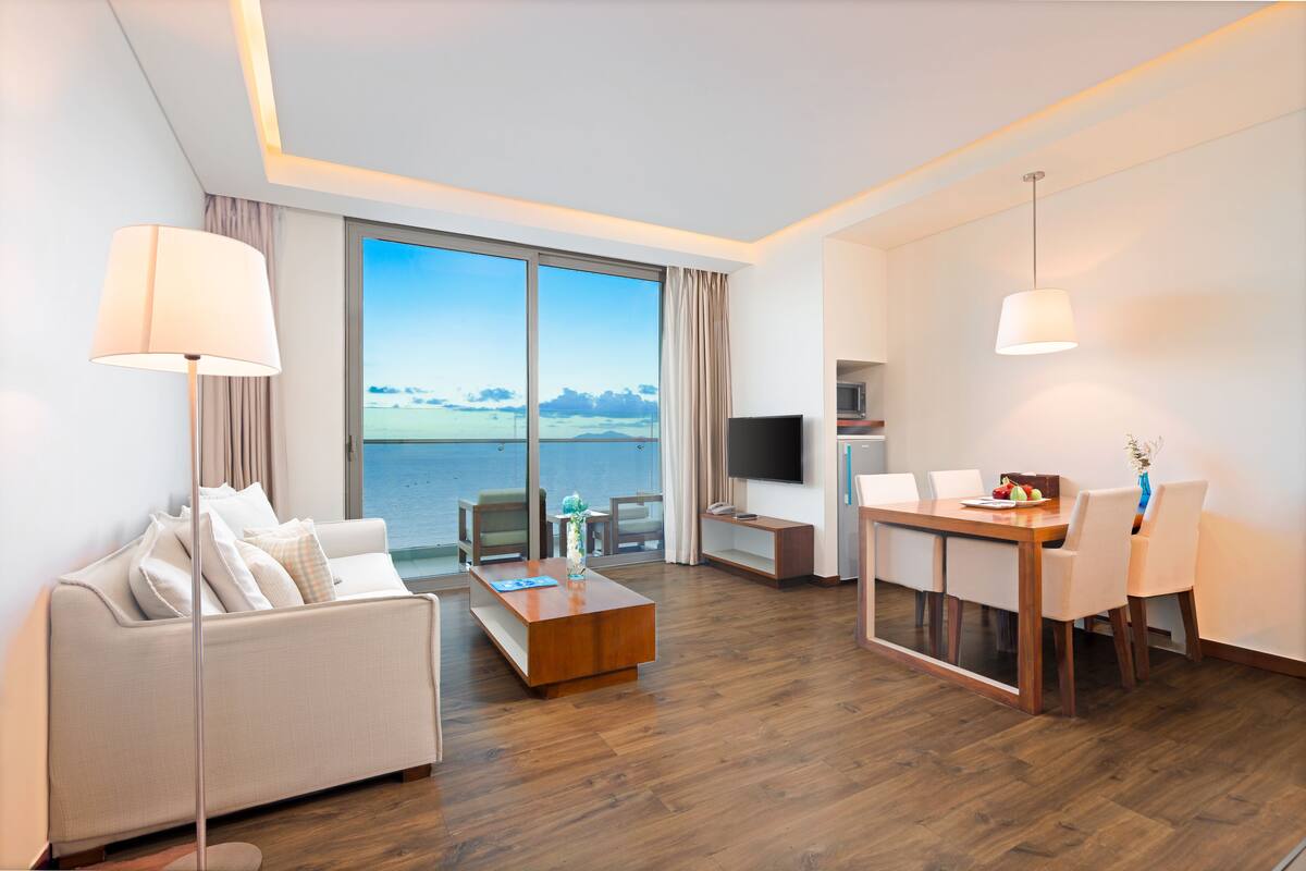 Une chambre d’hôtel moderne dotée d’une grande fenêtre, d’un canapé, d’une table basse, d’une table à manger avec des chaises, d’un téléviseur et d’une lampe.