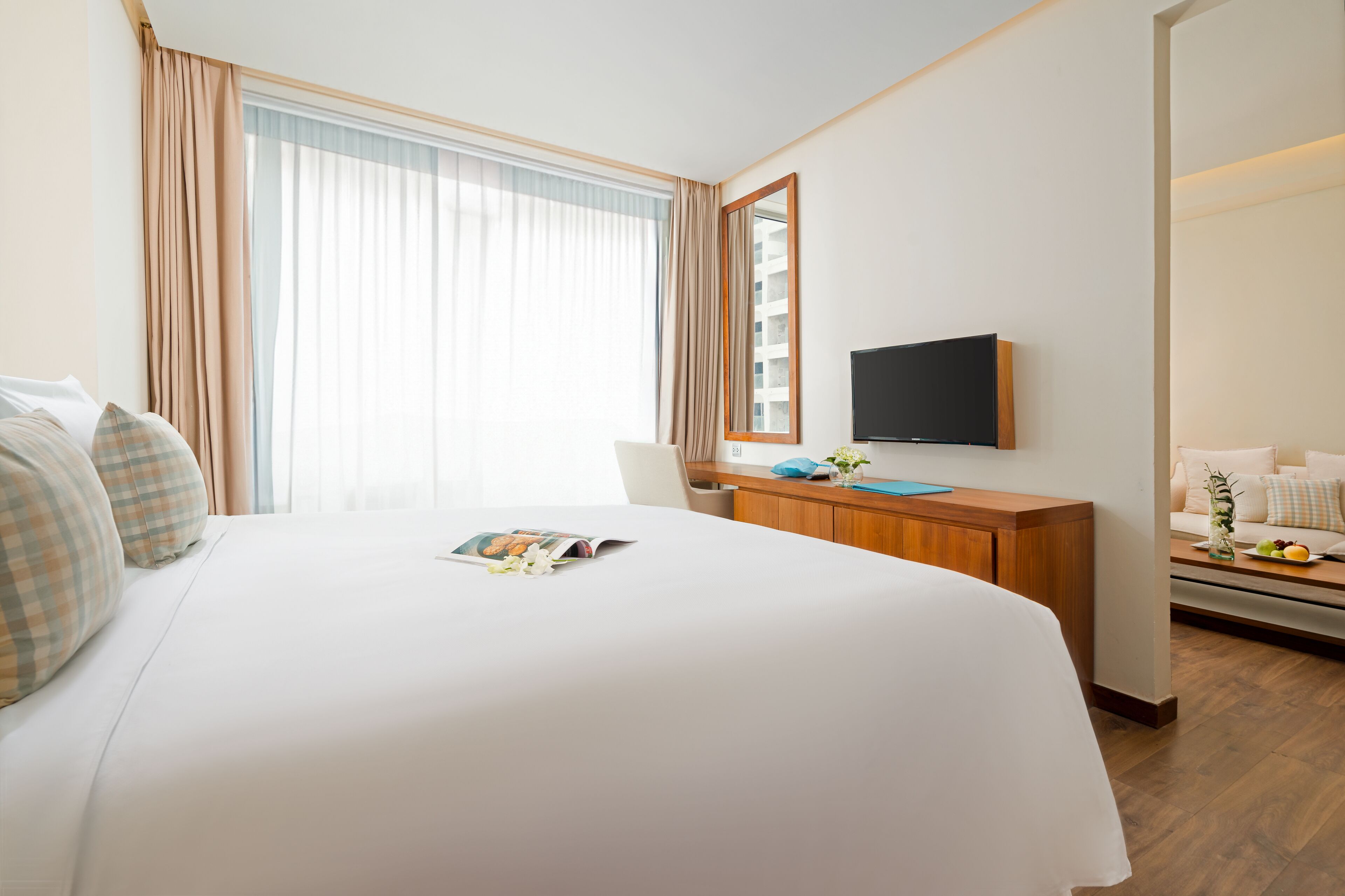 Ett hotellrum med en stor säng, ett skrivbord med en platt-tv, ett fönster med gardiner och ett litet bord med en vas och frukt.