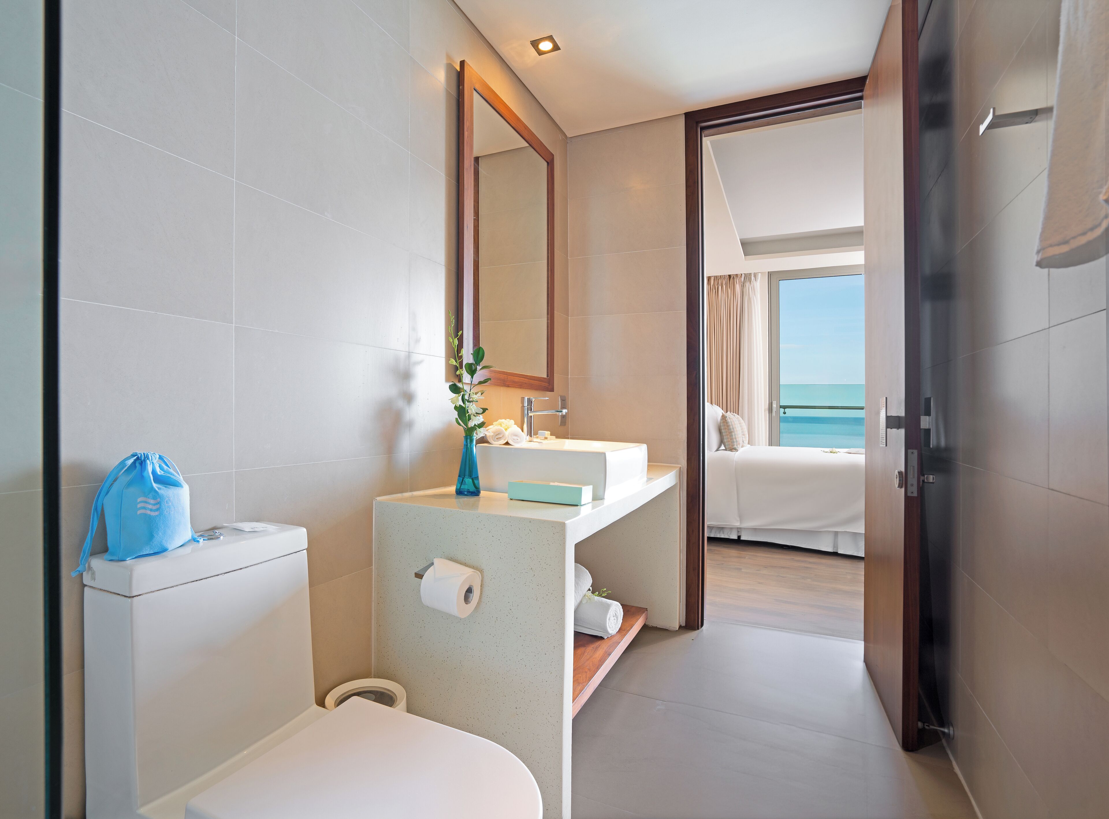 A modern bathroom with a sink, mirror, and toilet. A blue bag is on the counter, and a vase with flowers is on the sink.