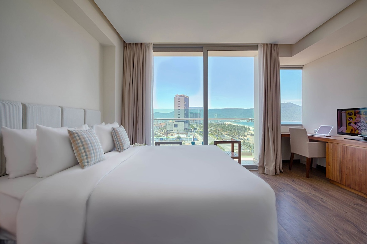 A hotel room with a large bed, a desk with a laptop, and a view of a cityscape through large windows.