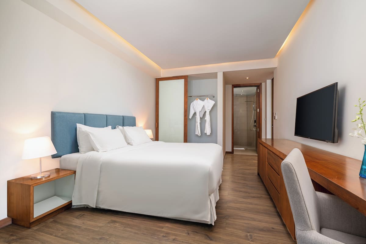 Une chambre d’hôtel moderne dotée d’un grand lit, d’une table de chevet avec une lampe, d’une commode en bois, d’un téléviseur fixé au mur et d’un coin salon avec un fauteuil.