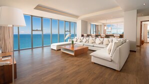 A spacious living room with a large window offering a view of the sea, a modern sofa set, a wooden coffee table, and a desk with a lamp.