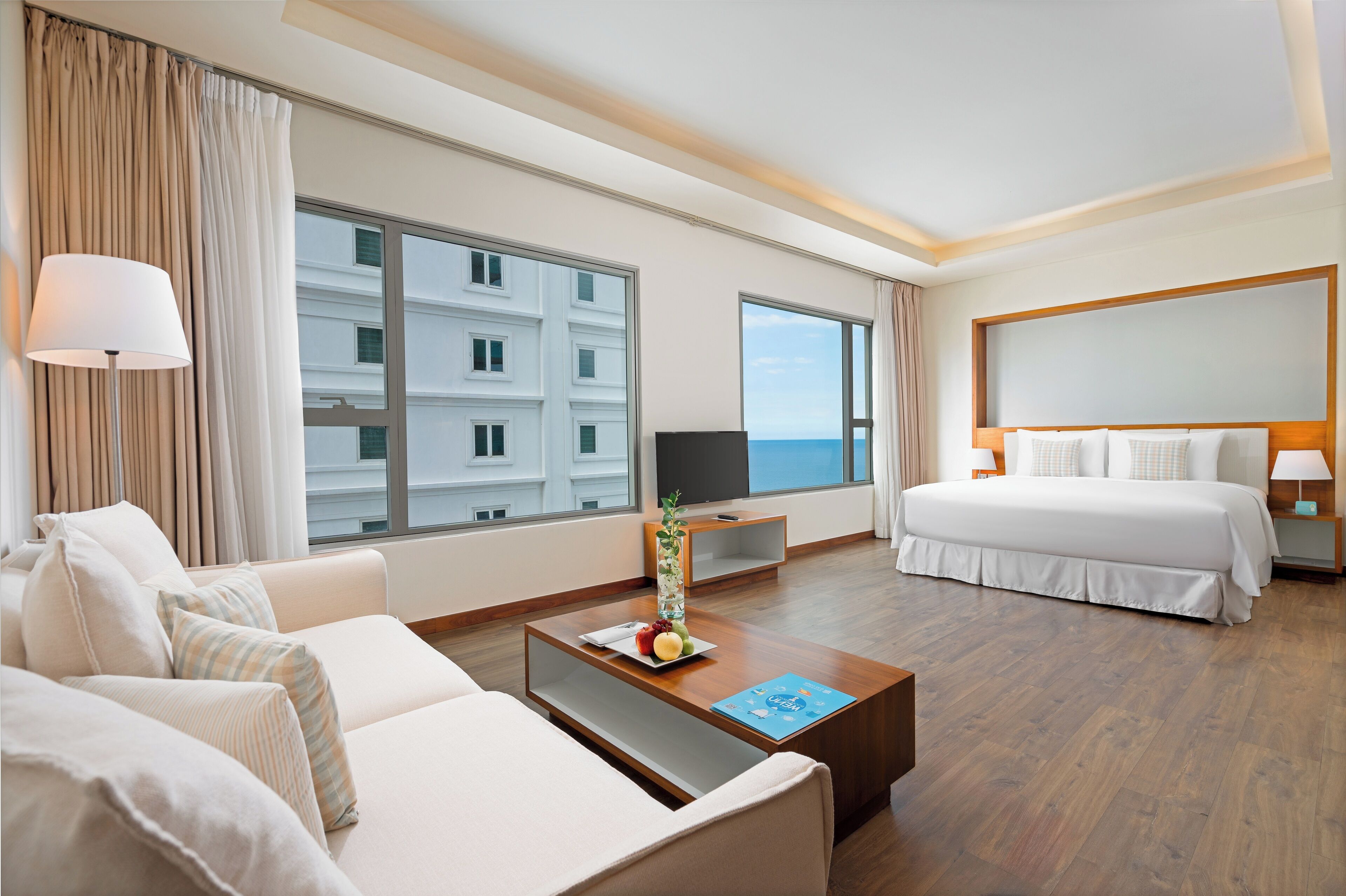 Une chambre d’hôtel avec une grande fenêtre, un lit, un canapé, une table basse avec des fruits et un téléviseur.