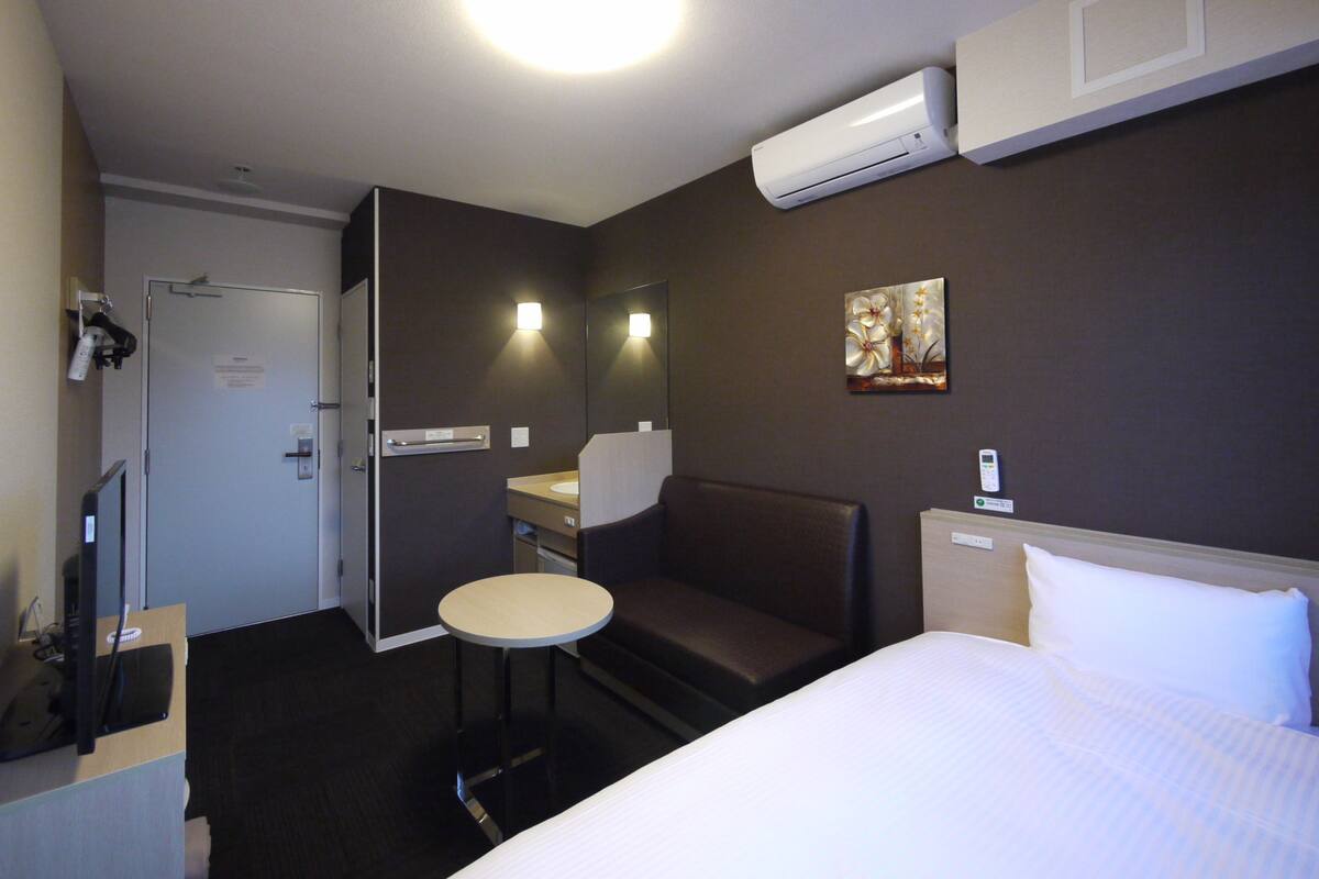 Hotellihuone, jossa on sänky, pieni pyöreä pöytä, tuoli, tietokoneella varustettu työpöytä, peili, ilmastointilaite ja taulu seinällä.