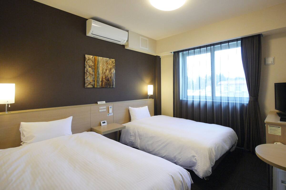 Hotellihuone, jossa on kaksi sänkyä, yöpöytä, työpöytä, televisio ja ikkuna, jossa on verhot.