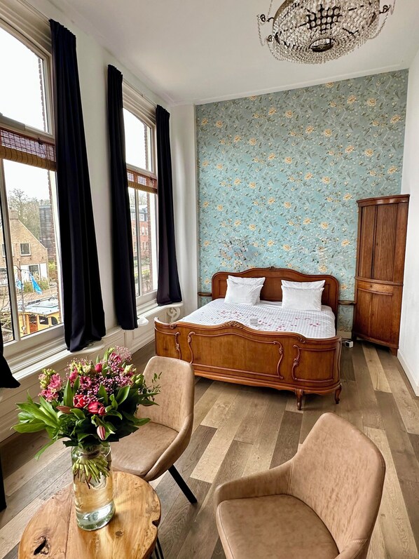 Une chambre avec un lit en bois, une armoire en bois, un vase de fleurs et une vue sur les bâtiments par la fenêtre.