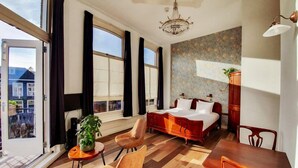 Une chambre d’hôtel moderne avec un grand lit, une table à manger et des chaises. On y trouve des fenêtres avec des rideaux, un lustre et une plante en pot.