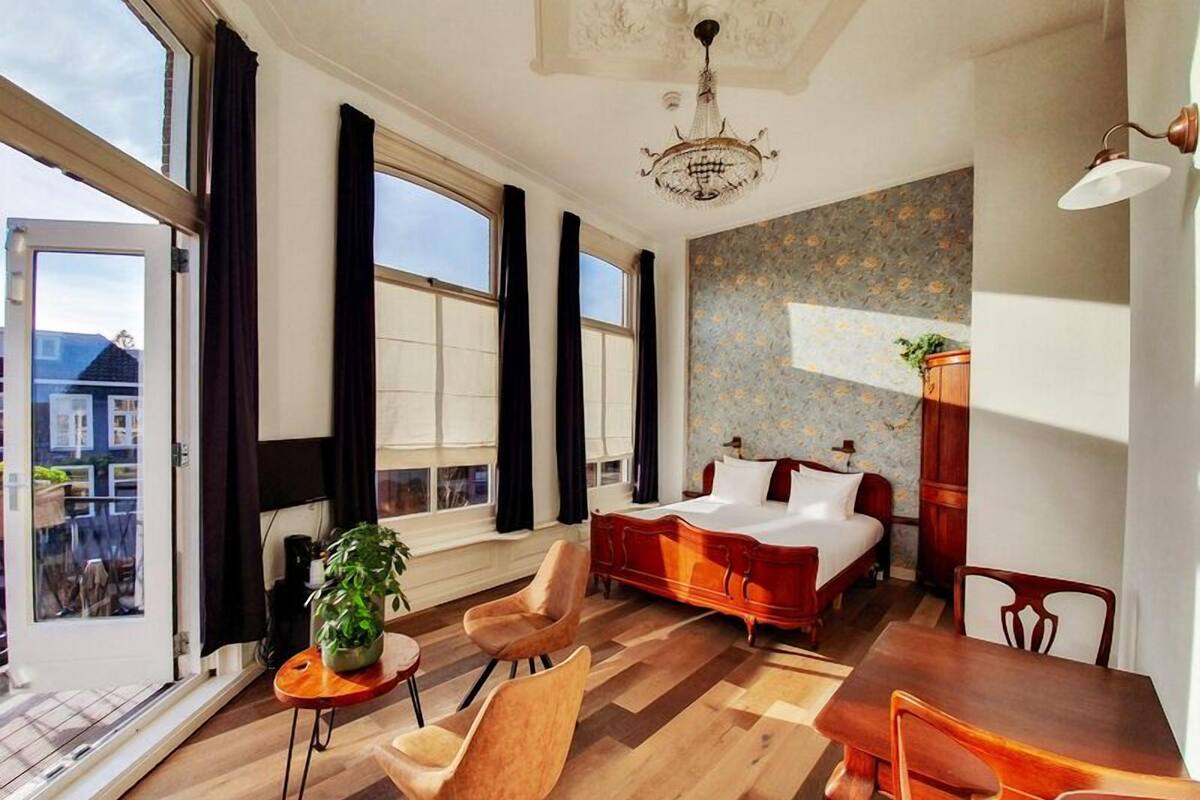 Une chambre d’hôtel moderne avec un grand lit, une table à manger et des chaises. On y trouve des fenêtres avec des rideaux, un lustre et une plante en pot.