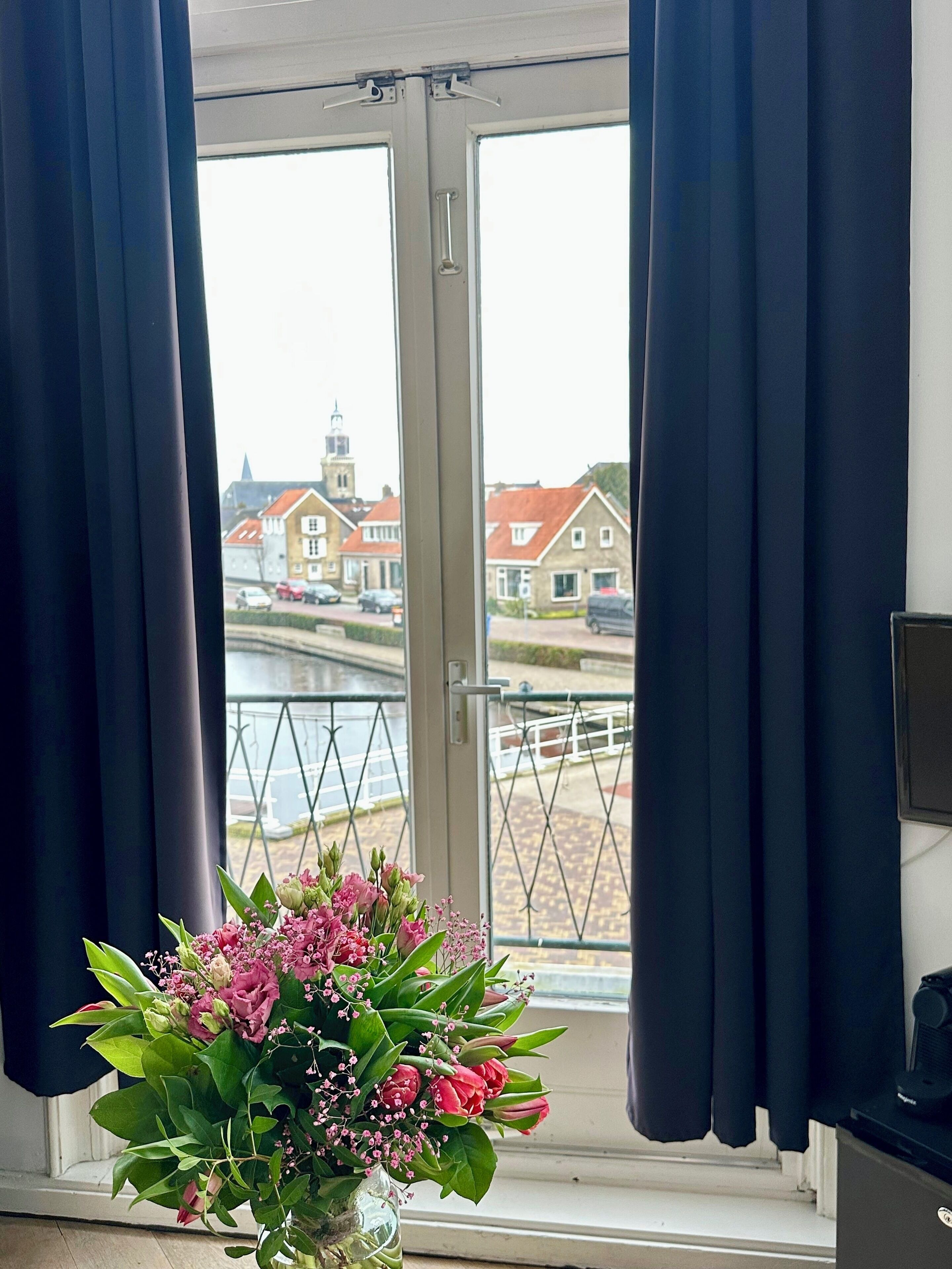 Una habitación con vistas a un canal y edificios, una ventana con cortinas oscuras y un florero con flores rosadas en el alféizar.