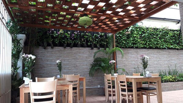 Un patio al aire libre con mesas y sillas de madera, un muro de piedra y diversas plantas en macetas.