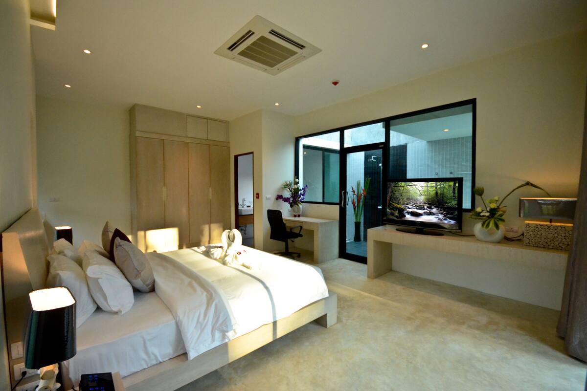 A modern hotel room with a large bed, a desk, a television, and a view of the outdoors through a glass door.