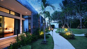 A modern house with a glass exterior, a well-maintained garden, and a pathway leading to the entrance. - Wanaburi Hotel (Thep Kasattri)