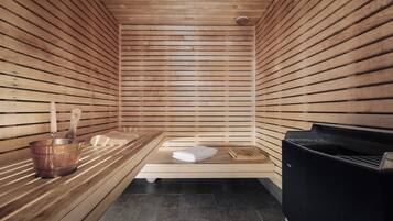A sauna with wooden slats, a bench, a towel, a wooden bucket, and a black heater.