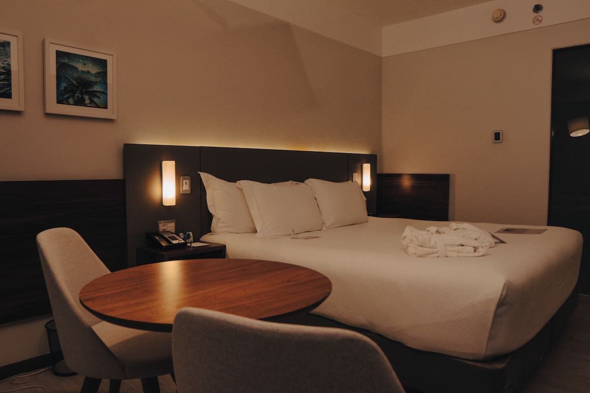 A hotel room with a bed, bedside table, lamp, and a round table with chairs.