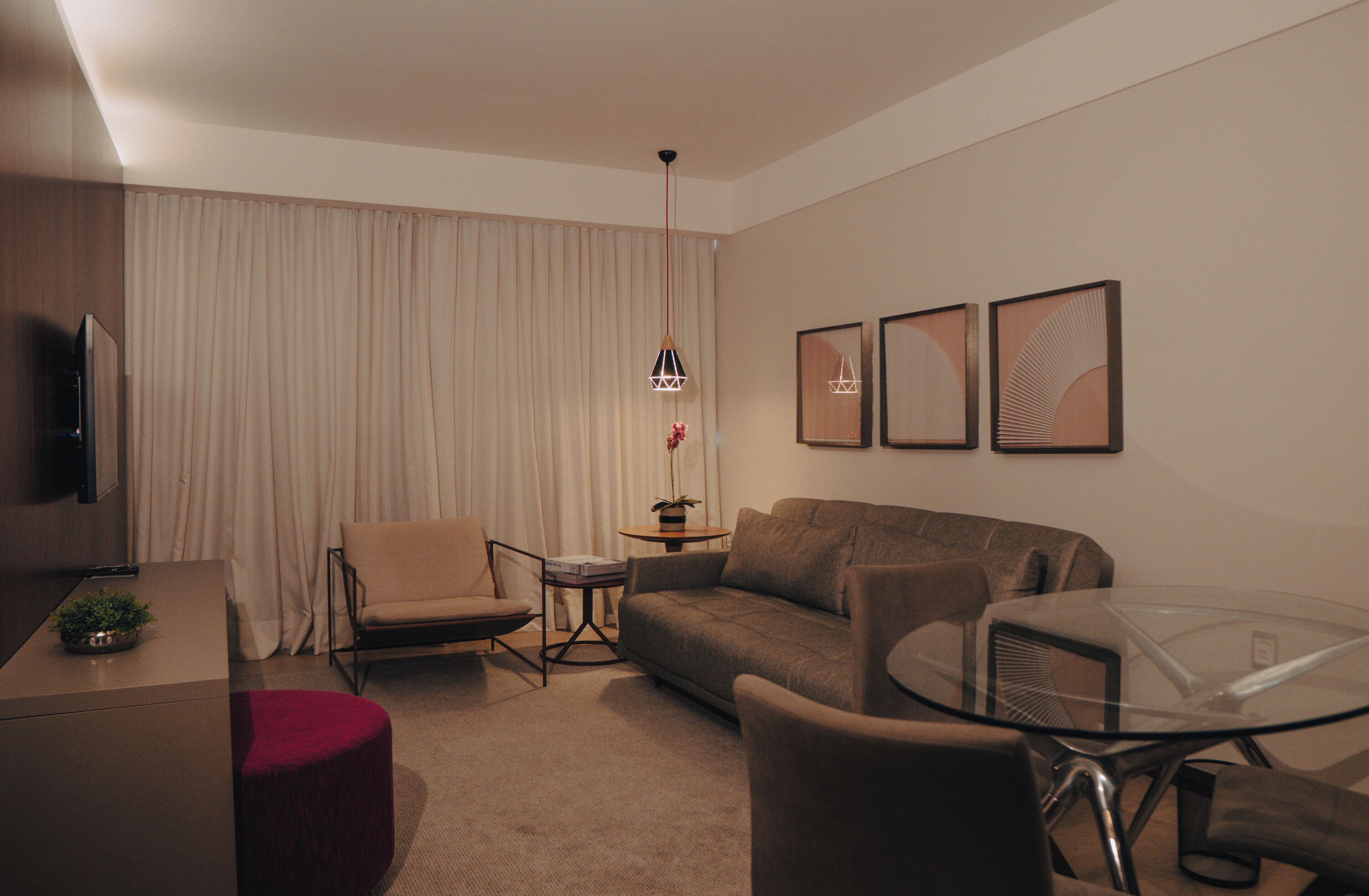 A modern hotel room with a sofa, armchairs, a glass coffee table, a TV, and a small side table with a plant.