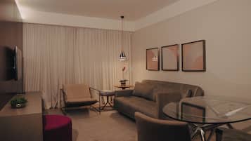 A modern hotel room with a sofa, armchairs, a glass coffee table, a TV, and a small side table with a plant.