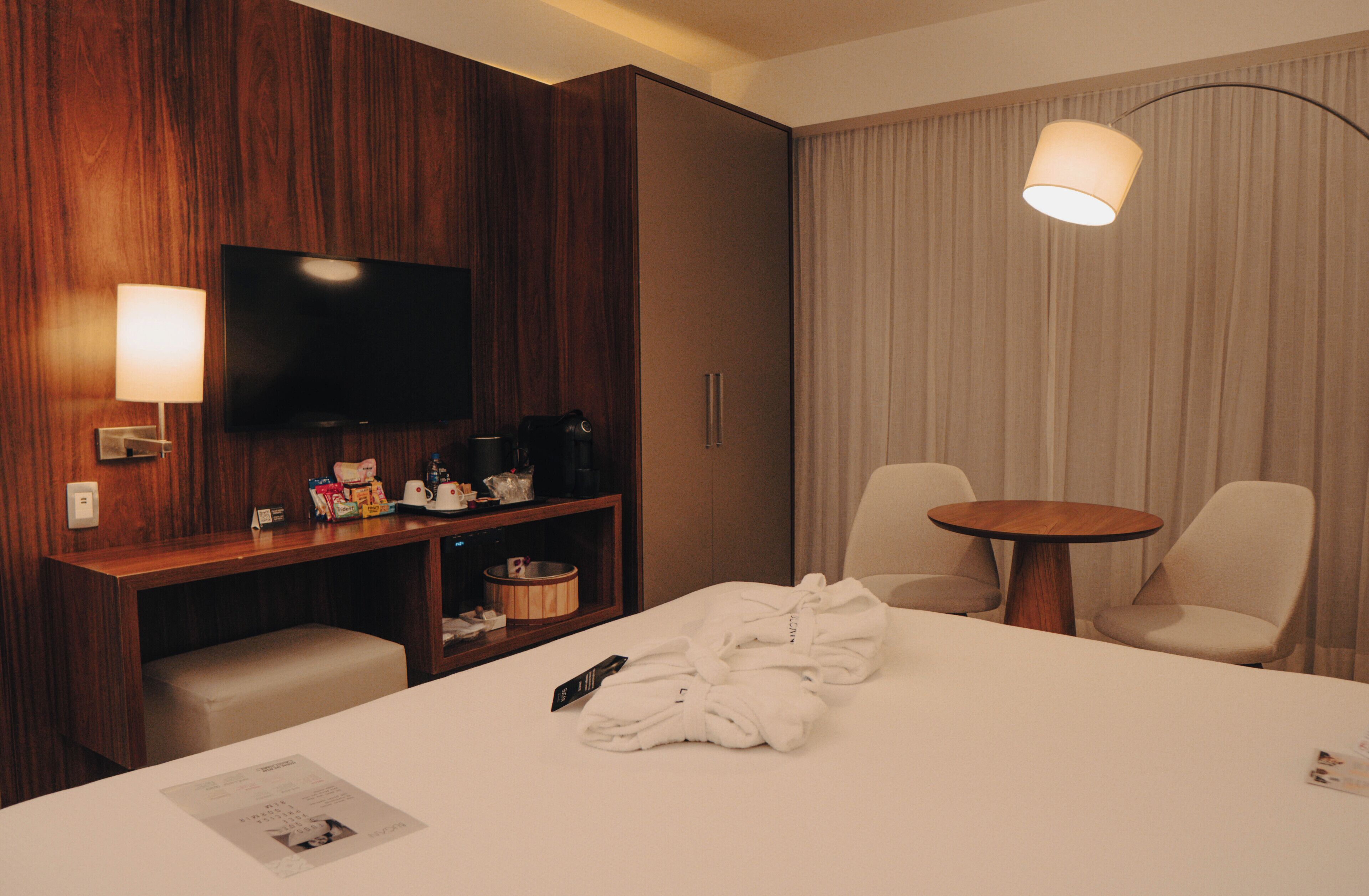 A hotel room with a bed, a TV, a desk with a lamp, a coffee machine, and a round table with chairs.