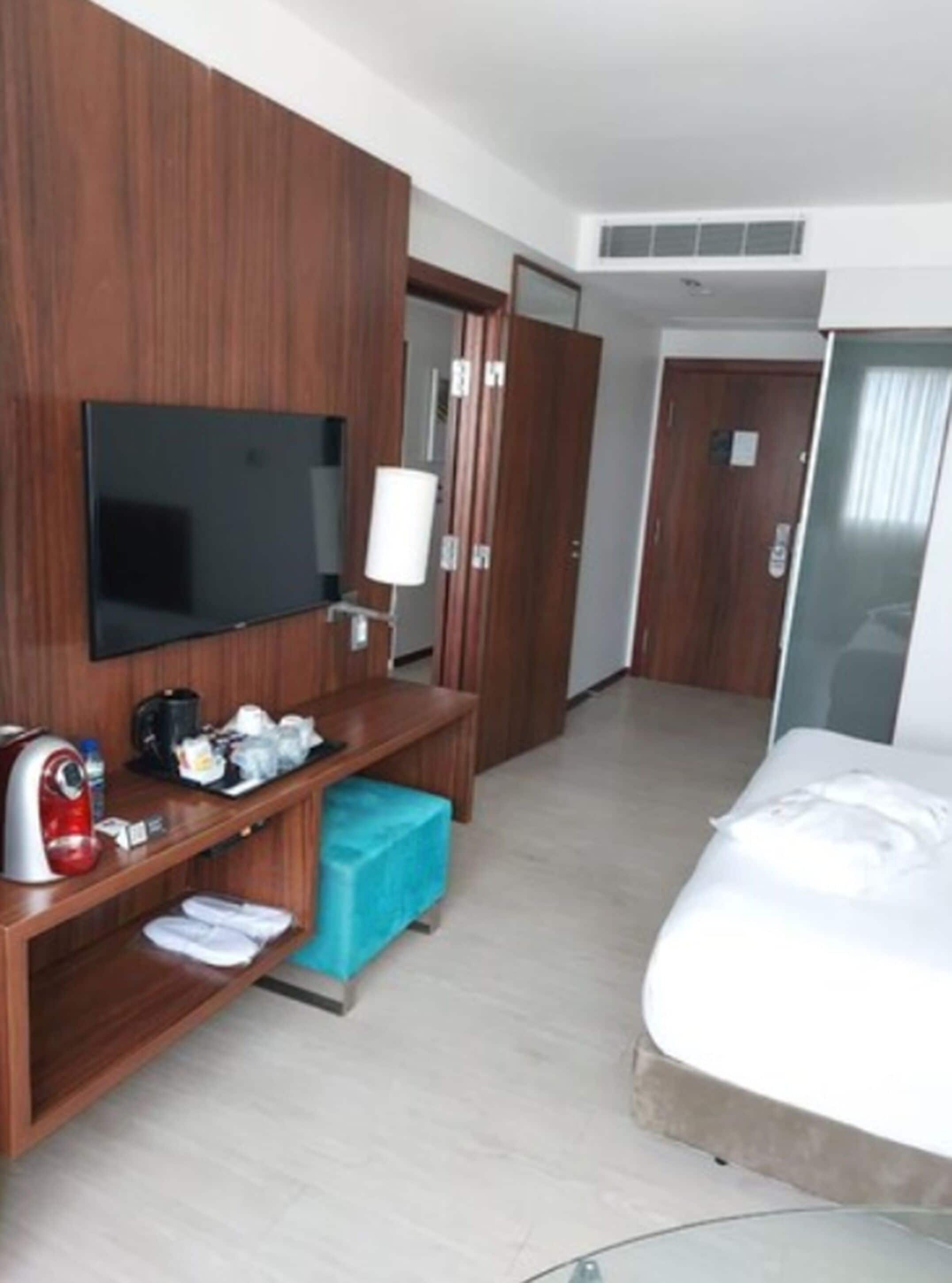 A hotel room with a bed, a TV mounted on the wall, a coffee machine, a small table, and a blue trash bin.