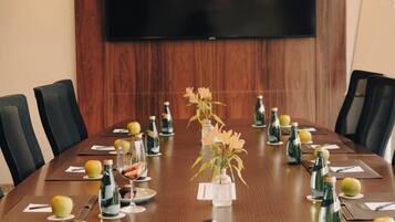 Uma sala de conferências com uma mesa comprida, cadeiras, uma televisão de tela grande e diversos objetos como garrafas e flores.