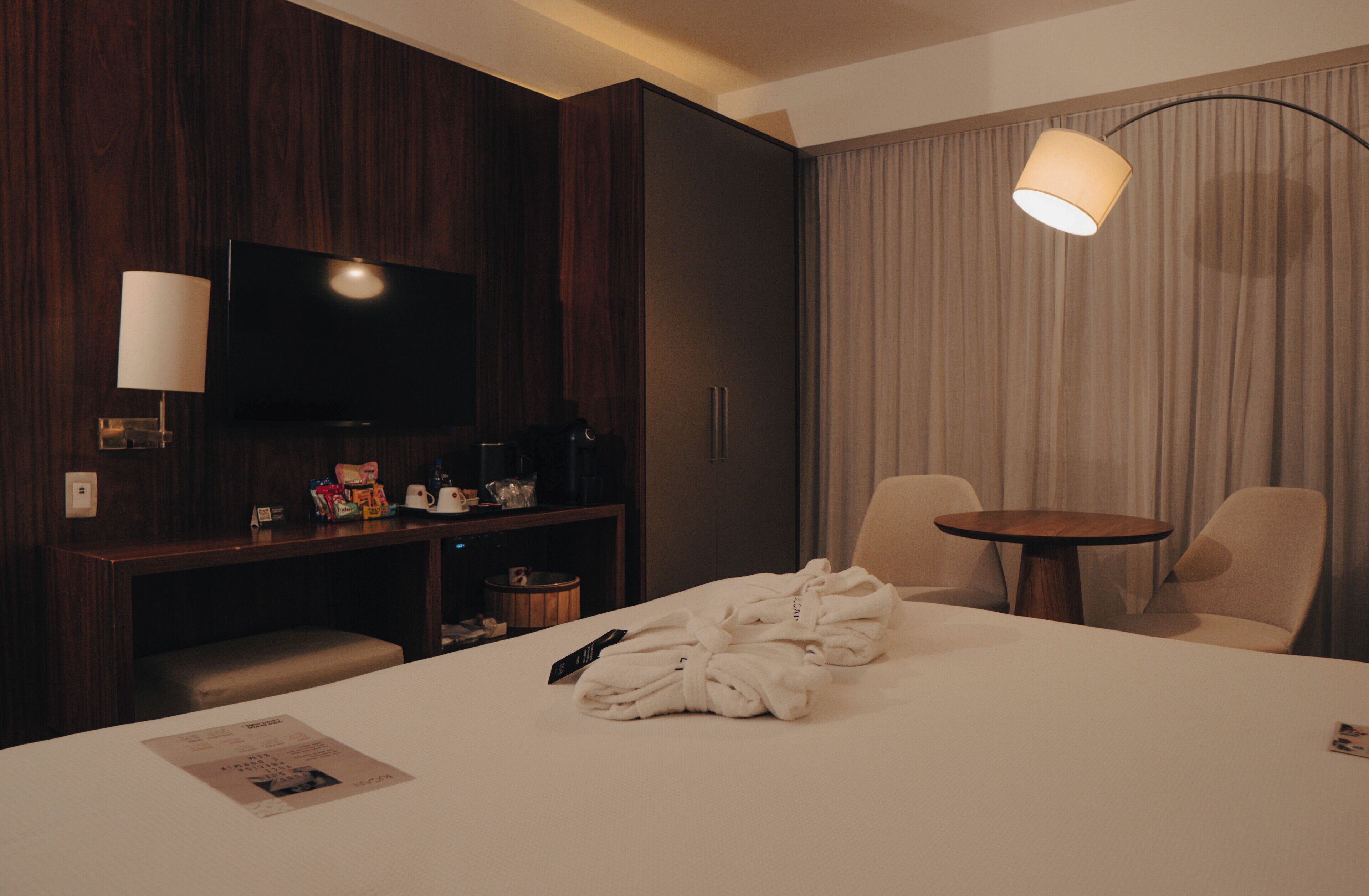 A hotel room with a bed, a TV, a desk with a lamp, a chair, and a table.