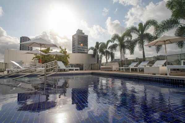 Una piscina con sillas de descanso y sombrillas, rodeada de palmeras y edificios modernos.