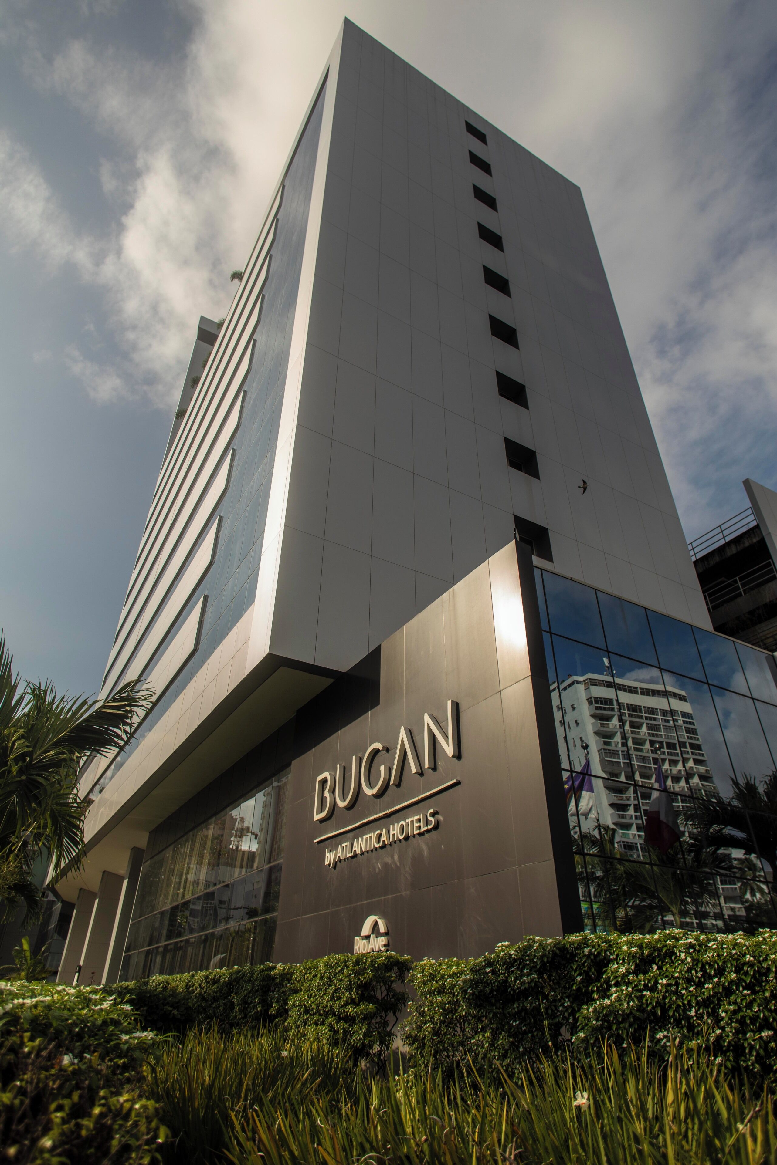 A modern hotel building with a prominent 'Bugan' sign, large windows, and a reflection of another building.