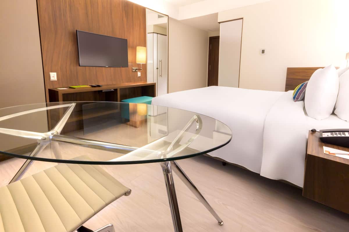 A modern hotel room with a glass table, a bed with white linens, a television mounted on the wall, and a wooden wardrobe.