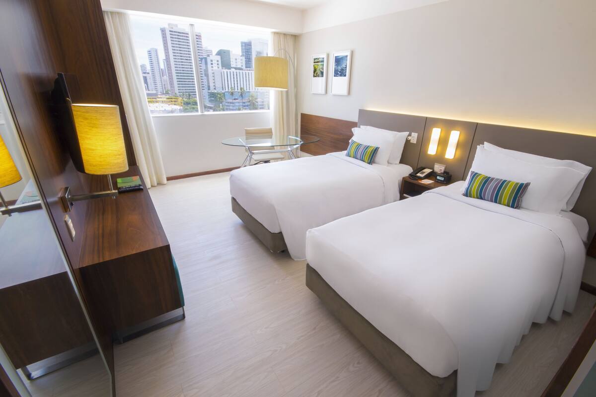 A hotel room with two beds, a desk, a chair, a lamp, a nightstand, and a window with a city view.