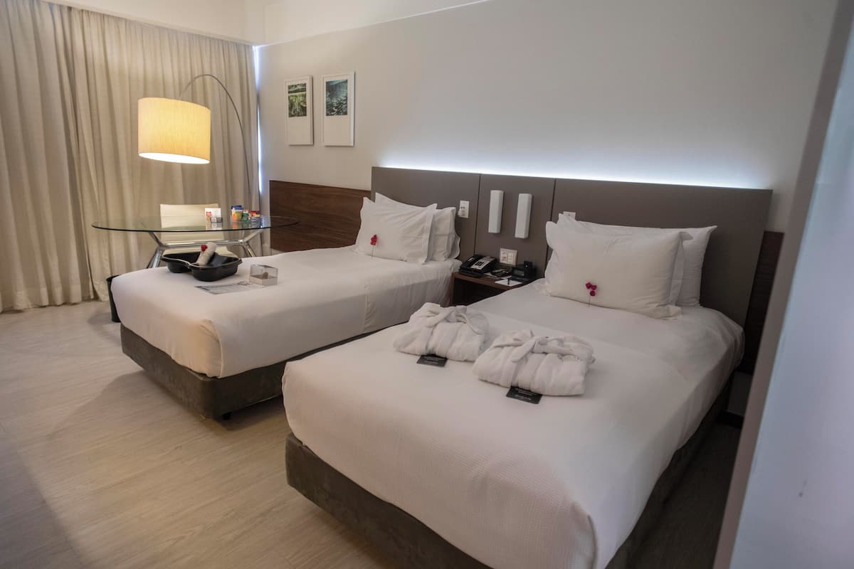 A hotel room with two beds, a glass side table, a lamp, and a nightstand.