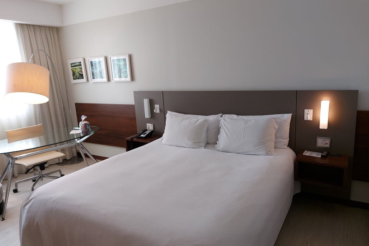 A hotel room with a large bed, a desk with a chair, a lamp, and a nightstand with a phone and a remote control.