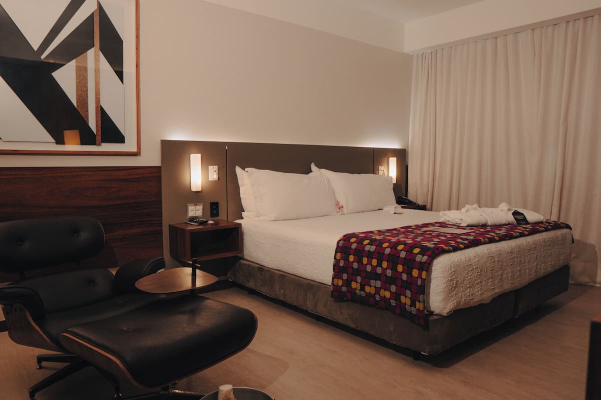 A hotel room with a bed, a desk, a chair, a wall-mounted TV, and a painting.