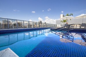 Una piscina en la azotea con baldosas azules cristalinas, barandilla de vidrio y vista a la ciudad.