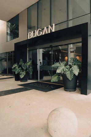 Un edificio con el letrero 'BUGAN', plantas en macetas grandes y una entrada de vidrio.