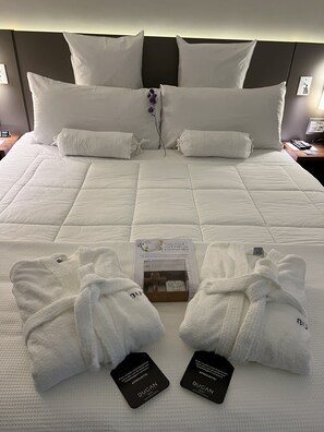 Una cama bien hecha con ropa blanca y almohadas, una sola flor morada y dos batas blancas con etiquetas.