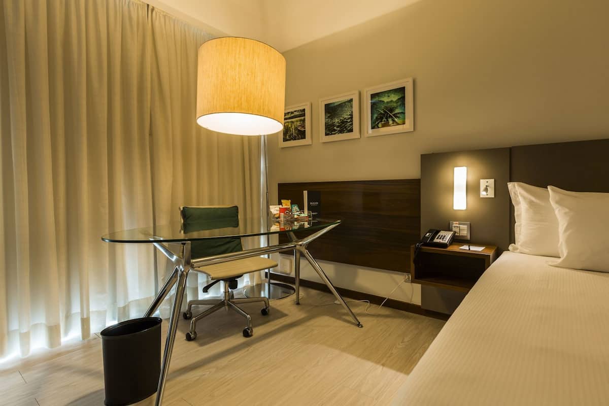 A hotel room with a bed, a desk with a chair, a lamp, a nightstand, and a wall with framed pictures.
