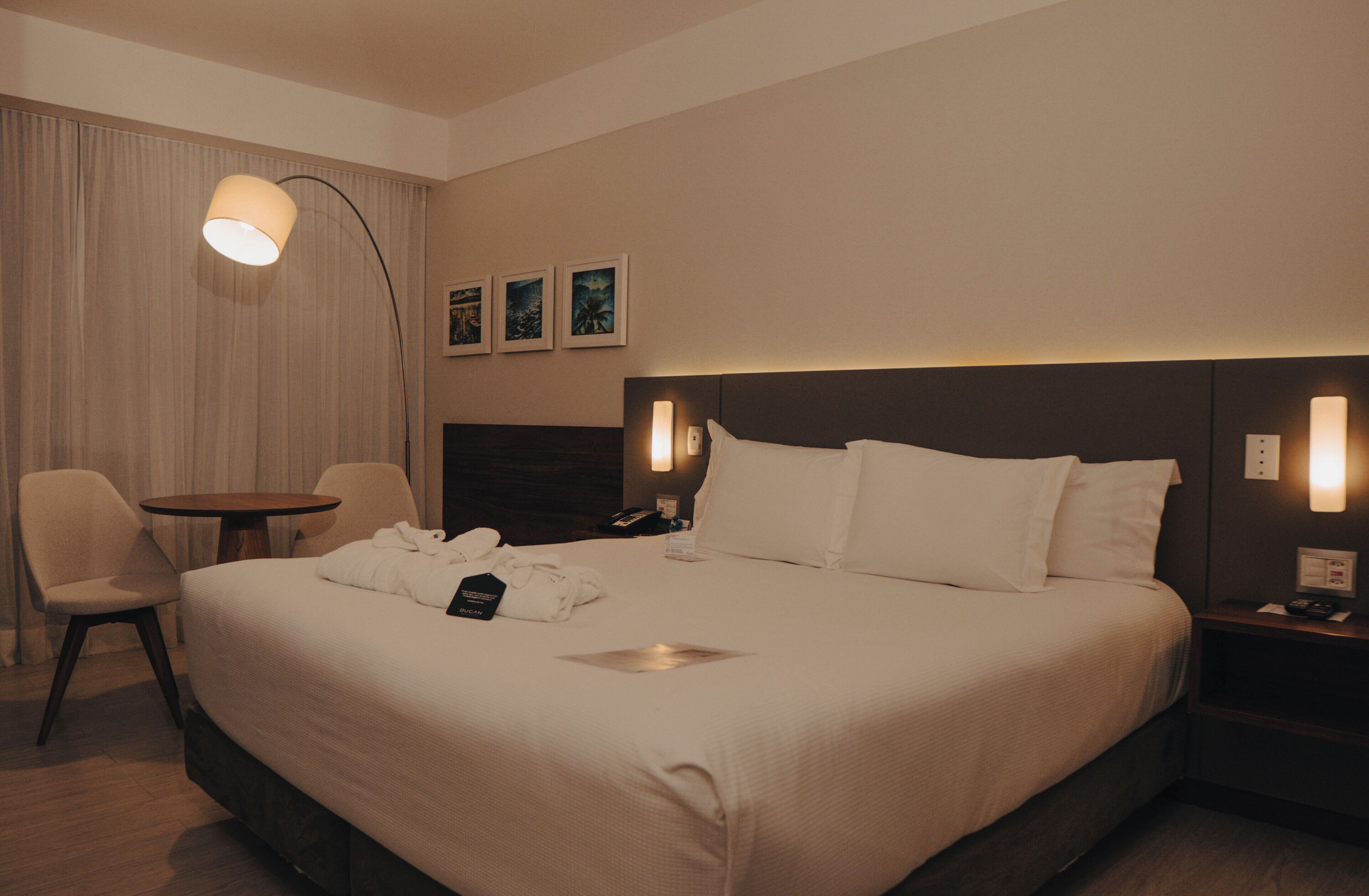 A hotel room with a large bed, a bedside table, a chair, a desk, and a lamp.