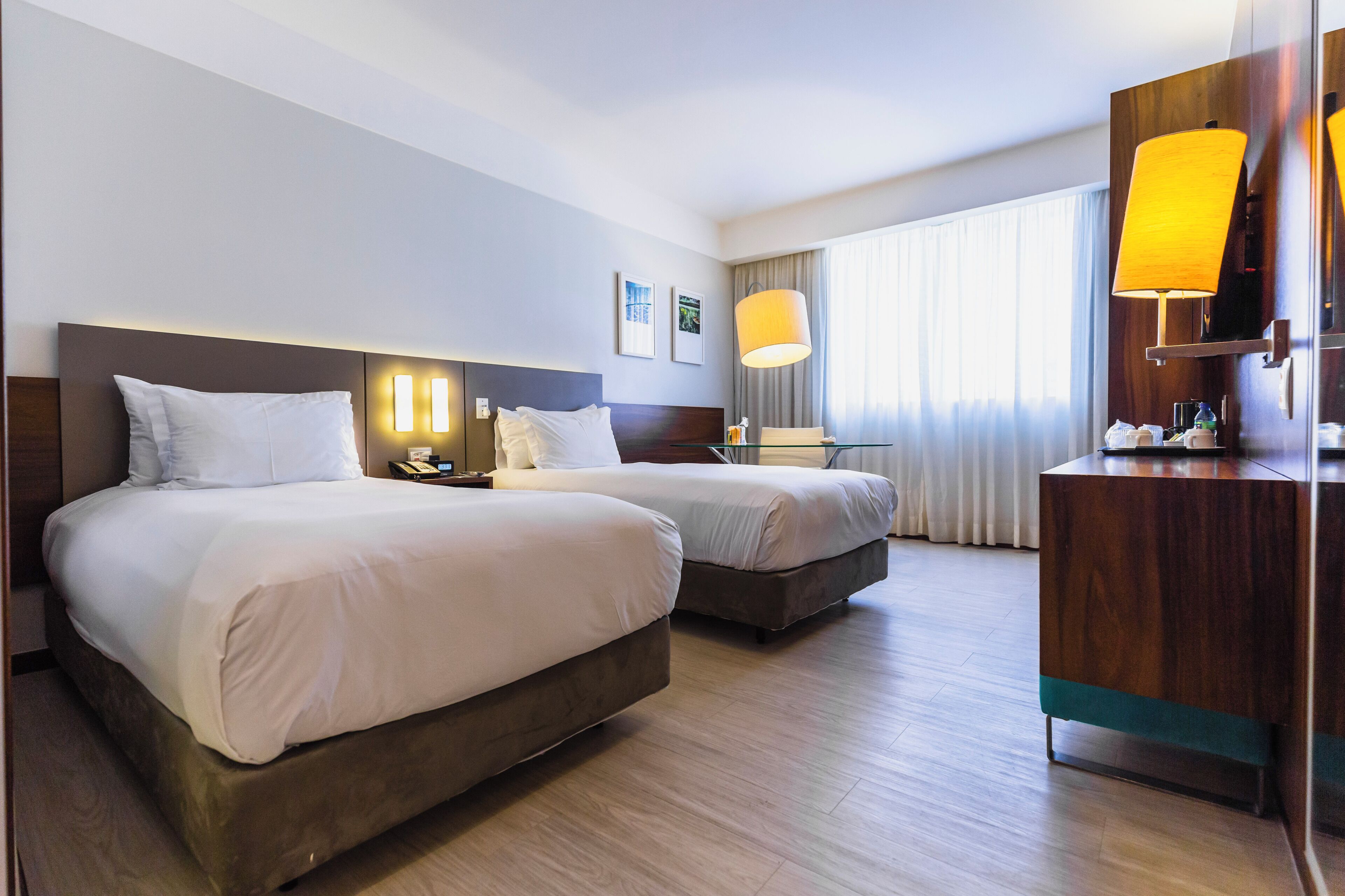 Habitación de hotel con dos camas, un escritorio de madera, una lámpara y un ventanal con cortinas.