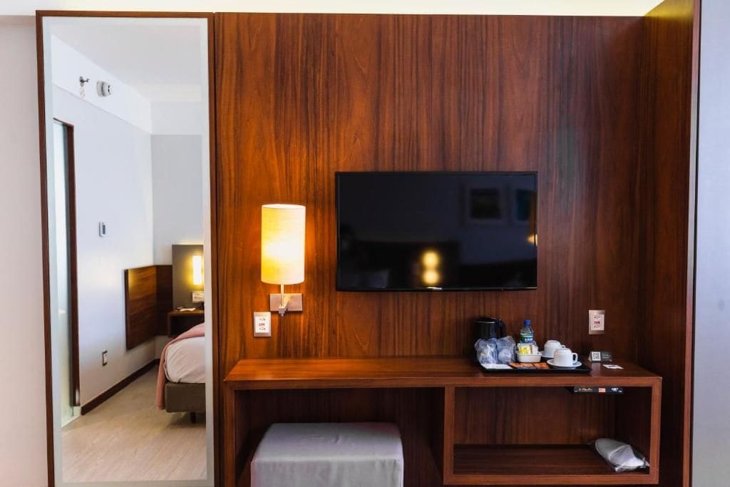 A hotel room with a wooden panel wall, a flat-screen TV mounted above a wooden console table, a bedside lamp, and a bed with a headboard.